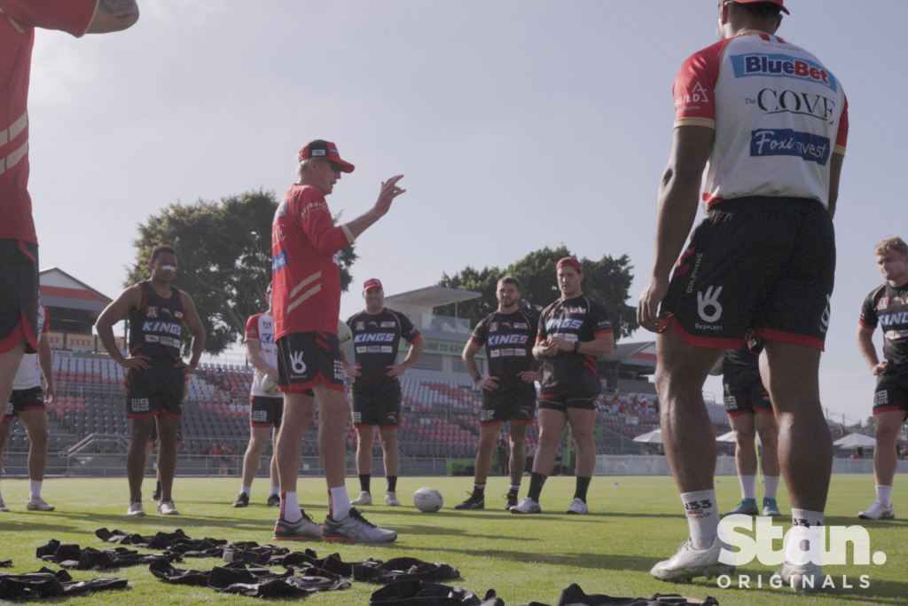 In a still shot from the new Stan TCV series Dawn of the Dolphins, supercoach Wayne Bennett speaks to players during a practice session. Bennett is wearing the Dolphins' jersey and the player stand in a circle around him in their training gear.