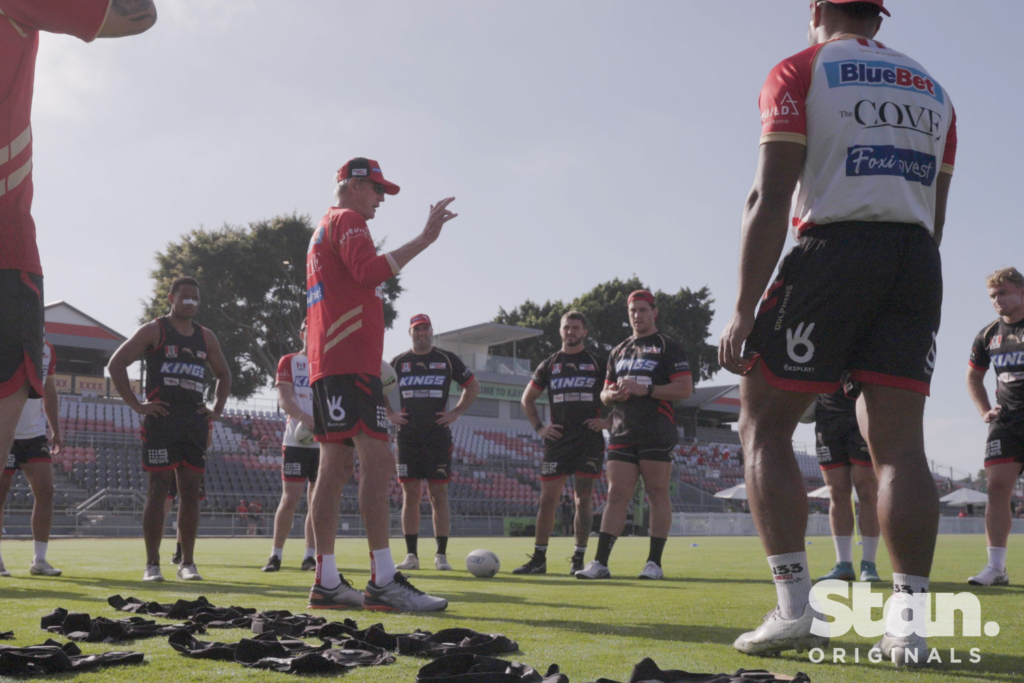 In a still shot from the new Stan TCV series Dawn of the Dolphins, supercoach Wayne Bennett speaks to players during a practice session. Bennett is wearing the Dolphins' jersey and the player stand in a circle around him in their training gear. 