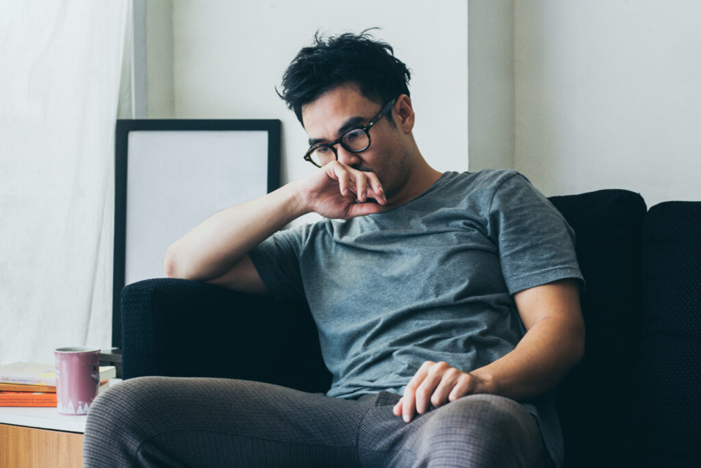 A man wearing glasses and a green T-shirt sits on a lounge chair looking downcast, with his hand covering his mouth.